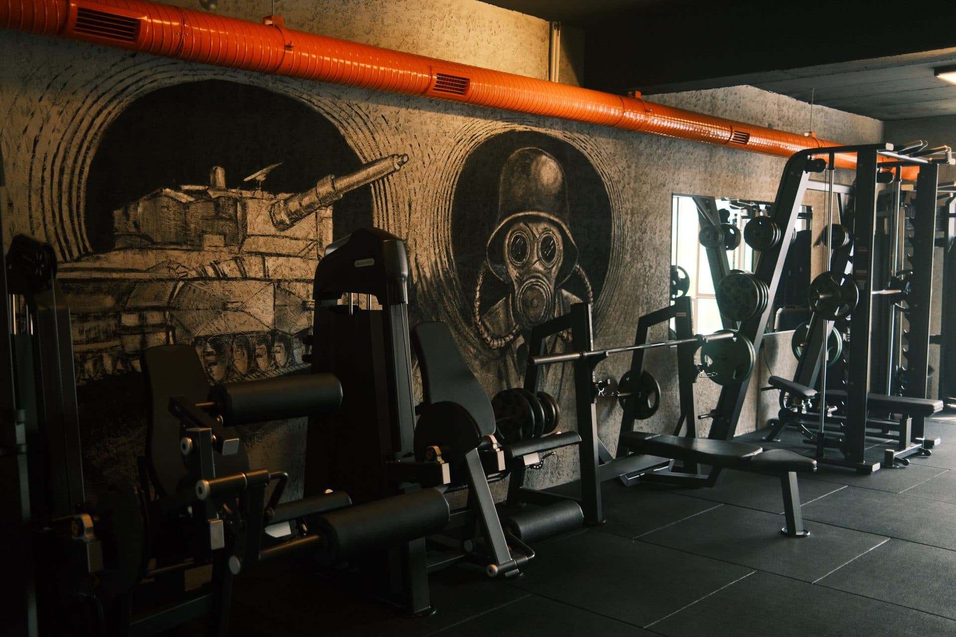 Karakalem tank ve gaz maskeli figür muralleri önünde bacak ekstansiyon-curl makineleri ve bench